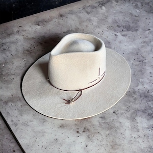 May include: A cream-colored cowboy hat with a wide brim and a rounded crown. The hat features a brown leather band and decorative stitching. A leather cord with decorative beads hangs from the hat.