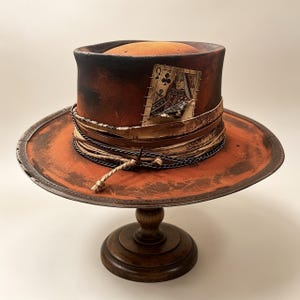 May include: A distressed brown top hat featuring a Queen of Clubs playing card stitched onto the side. The hat is adorned with layered leather bands, a thin dark metal chain, and a piece of twine, all set against a weathered, burnt orange finish.