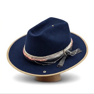May include: A navy blue fedora hat with a wide brim and decorative trim. The brim has a tan edge. The hat is adorned with a band of silver and pink strands, and a dark grey ribbon. The hat is displayed on a wooden stand.