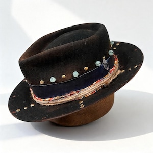 Könnte beinhalten: Dunkelbrauner Fedora-Hut mit breiter Krempe. Der Hut hat ein marineblaues Band, dekorative goldene Nieten und türkisfarbene Perlen. Ein gemusterter Stoff und eine Kordel verzieren das Hutband. Der Hut steht auf einem braunen Hutständer.