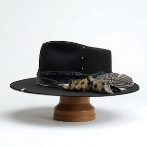 May include: A black fedora hat with a wide brim, featuring a decorative band, feathers, and a leopard print accent. The hat is presented on a wooden stand, highlighting its design and style.