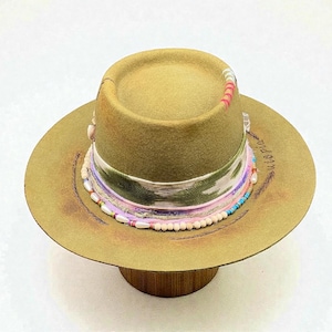 Pode incluir: Um chapéu fedora bege com aba larga e uma faixa decorativa. A faixa apresenta um tecido estampado verde e branco, fitas rosa e roxas e uma fileira de contas brancas. A palavra "utopia" está escrita na aba.