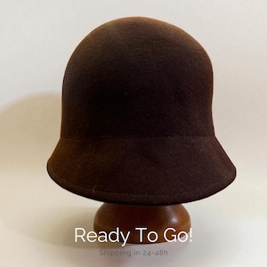 Può includere: Un cappello cloche marrone scuro con una corona arrotondata e una tesa leggermente svasata. Il cappello è realizzato in un materiale morbido simile al feltro ed è esposto su un espositore per cappelli in legno. Il testo "Ready To Go!" è visibile.
