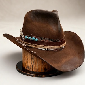 Könnte beinhalten: Ein brauner Cowboyhut mit einem dekorativen Band mit türkisfarbenen Perlen und einer Feder. Der Hut wird auf einer Holzspule präsentiert. Der Hut hat eine breite Krempe und eine klassische Cowboyhutform.