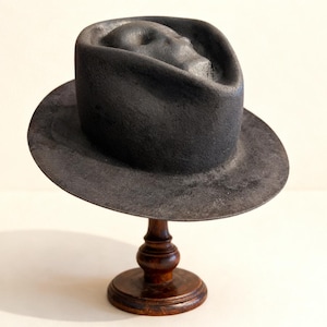 Può includere: Un cappello fedora grigio scuro con un'indentazione a forma di volto sulla corona, esposto su un supporto di legno. Il cappello ha un'ampia tesa e una superficie strutturata. Il supporto è in legno scuro con base rotonda.