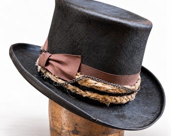 May include: A black top hat featuring a brown ribbon band with a bow, a braided beige fabric trim, and a thin dark metal chain wrapped around the crown. The hat has a wide, slightly curved brim and a textured, matte black finish.