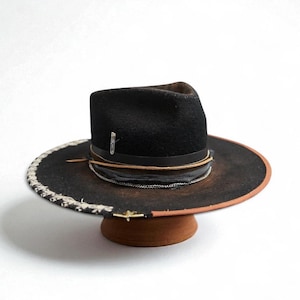 Könnte beinhalten: Schwarzer Fedora-Hut mit breiter Krempe, mit dekorativem Band und Kristallnadel. Der Rand hat einen verwitterten braunen Rand und eine dekorative Verzierung. Der Hut wird auf einem braunen Hutständer präsentiert.