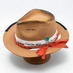May include: A tan fedora hat with a decorative band. The band features a white and black patterned fabric, an orange fabric accent, and a turquoise stone. The hat has a black band around the brim and a stitched design on the crown.