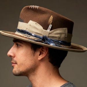 Könnte beinhalten: Ein brauner Fedora-Hut mit dekorativer Feder und Band. Der Hut hat ein hellbraunes Band, ein dunkelblaues Band und eine dekorative Feder. Der Hut hat eine breite Krempe und ein gesticktes Design auf der Krone.