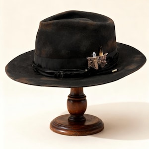 Peut inclure: Un chapeau fedora noir vieilli à large bord, orné d'un ruban noir et de décorations. Le chapeau est présenté sur un présentoir en bois, mettant en valeur son style vintage et son savoir-faire. Le bord du chapeau a un aspect usé.