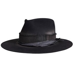 Könnte beinhalten: Ein schwarzer Fedora-Hut mit breiter Krempe und einem Ripsband. Der Hut hat ein leicht abgenutztes Aussehen mit weicher Textur. Die Krempe des Hutes misst etwa 7,6 cm.