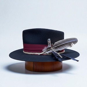 Könnte beinhalten: Schwarzer Fedora-Hut mit breiter Krempe, burgunderfarbenem Band und dekorativer Feder- und Kristallverzierung. Der Hut wird auf einem hölzernen Hutblock präsentiert, um seine Form und sein Design zu zeigen.