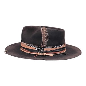 May include: A dark grey fedora hat with a wide brim. The hat features a patterned band, a leather band, and a feather accent. A rope is wrapped around the base of the hat. The hat has a distressed appearance.