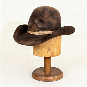 Può includere: Un cappello da cowboy marrone invecchiato con tesa larga, esposto su un espositore in legno. Il cappello presenta una fascia di corda attorno alla corona. L'espositore è in legno e ha una base rotonda.