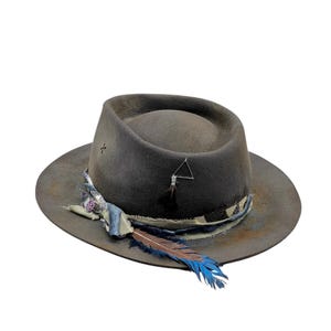 May include: A distressed grey fedora hat with a wide brim. The hat features a patterned fabric band, a feather, and a small silver triangle charm. The feather is brown and blue.