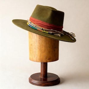 Puede incluir: Sombrero fedora verde oliva con ala ancha, adornado con una banda roja y un ribete tejido colorido. El sombrero se exhibe en un soporte de sombrero de madera. El sombrero tiene un diseño clásico y elegante.