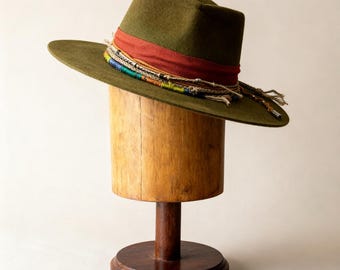 May include: An olive green fedora hat with a wide brim, featuring a red band and colourful woven trim. The hat is displayed on a wooden hat stand. The hat has a classic design. The hat stand is made of wood.
