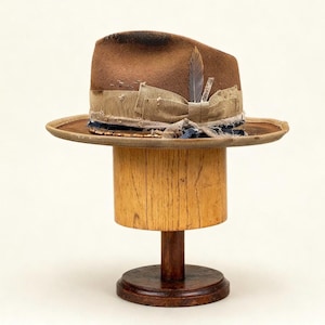 Könnte beinhalten: Ein brauner Fedora-Hut mit dekorativem Band und Federakzent. Der Hut wird auf einem hölzernen Hutständer mit rundem Sockel und zylindrischem Pfosten präsentiert. Der Hut hat eine breite Krempe.