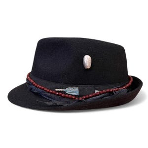Puede incluir: Un sombrero fedora negro con una banda decorativa de cuentas rojas y un adorno de pluma plateada. El sombrero presenta una pequeña piedra ovalada en el lateral. Un accesorio elegante.