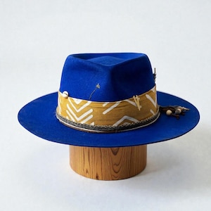 Peut inclure: Un chapeau fedora bleu vif avec un large bord. Le chapeau est orné d'un bandeau jaune moutarde avec un motif géométrique blanc. Il est orné d'un petit coquillage, d'herbe séchée et de perles en bois. Le chapeau est présenté sur un support en bois.