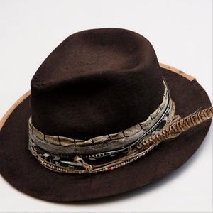 Könnte beinhalten: Ein dunkelbrauner Fedora-Hut mit dekorativem Band und Federakzent. Der Hut hat eine breite Krempe und ein gemustertes Band mit Perlen. Die Feder ist hellbraun.