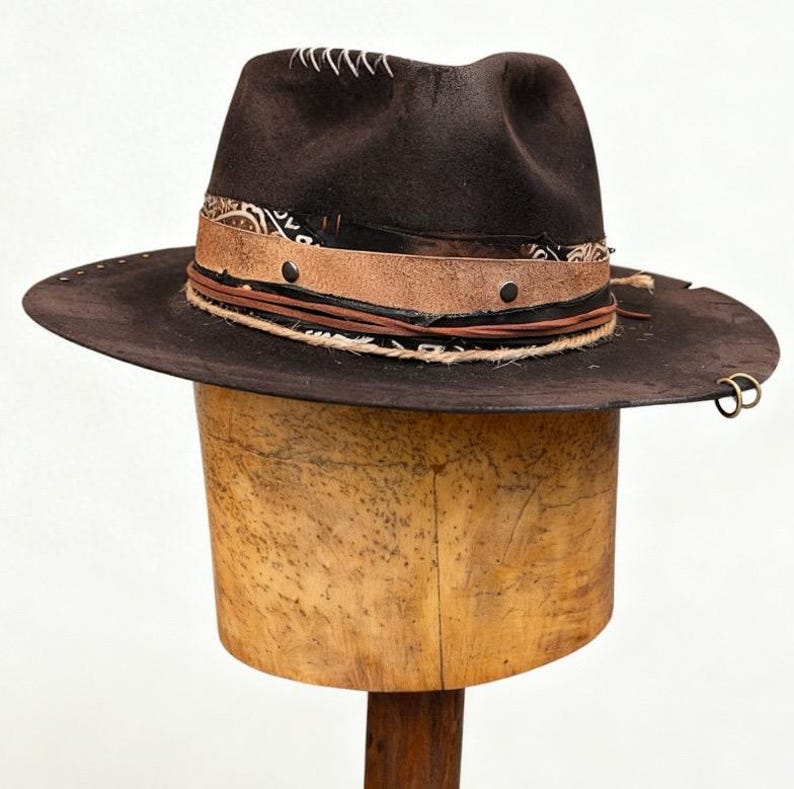 May include: A dark brown fedora hat with a wide brim. The hat has a decorative band with leather and rope detailing, and a patterned fabric. The hat is displayed on a wooden hat form.