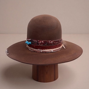 Puede incluir: Sombrero fedora marrón con ala ancha, adornado con una banda estampada en tonos rojos y marrones. El sombrero presenta una pluma y cuentas turquesas. Se exhibe sobre un soporte de madera.