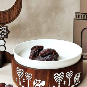 May include: A white ceramic bowl filled with dates sits atop a wooden serving tray. The tray is decorated with white palm trees and camels. Decorative wooden crescent moon and architectural elements are in the background.