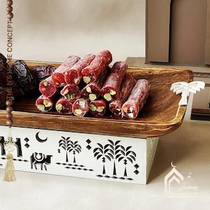 May include: A wooden serving tray filled with Turkish delight, a traditional Middle Eastern dessert. The red, cylindrical sweets are studded with pistachios. The tray sits atop a white decorative stand with palm tree cutouts. Dates are also visible.