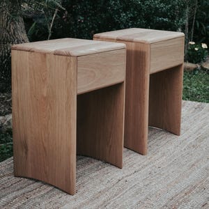 May include: Two wooden bedside tables with a single drawer each. The tables are made of light brown wood and have a curved base. The tables are sitting on a woven rug.