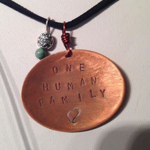 May include: A copper pendant necklace with a black cord. The pendant is round and has the words "ONE HUMAN FAMILY" engraved on it. A small heart is engraved below the text.