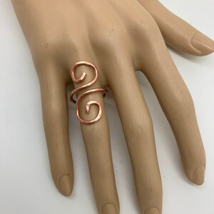 May include: A hammered copper ring with a swirling design. The ring is worn on a model's finger. "Hammered Copper 1x Wrap" is written in pink text at the bottom of the image.