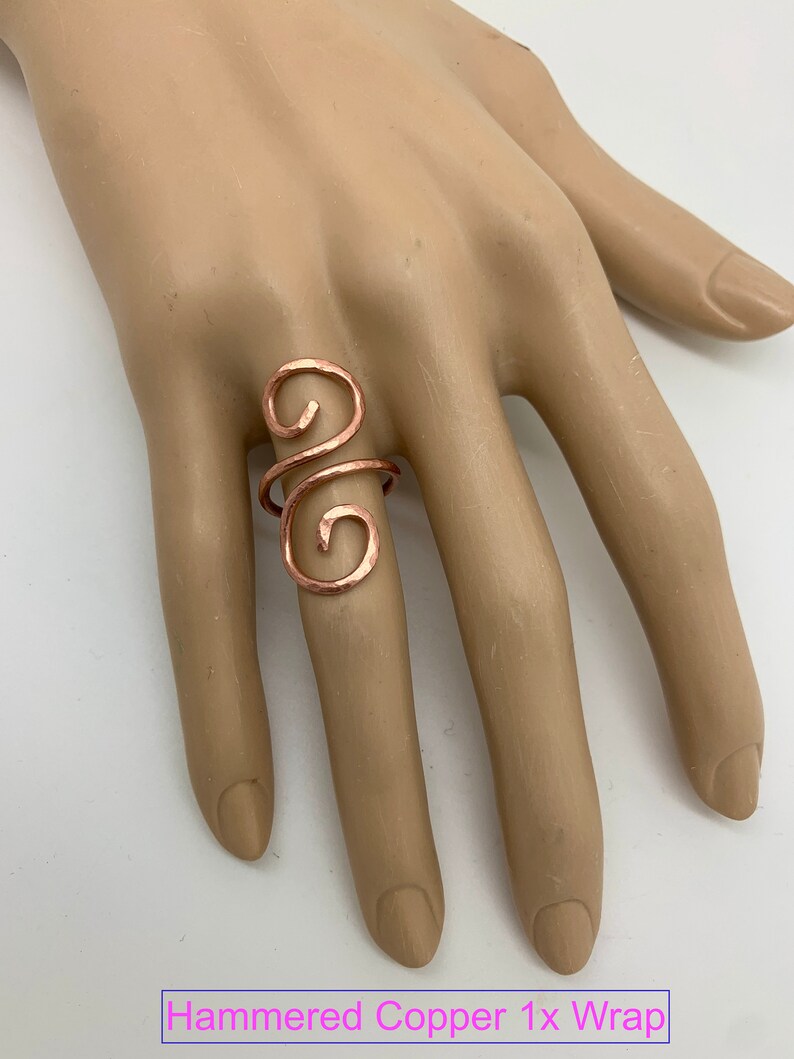 May include: A hammered copper ring with a swirling design. The ring is worn on a model's finger. "Hammered Copper 1x Wrap" is written in pink text at the bottom of the image.