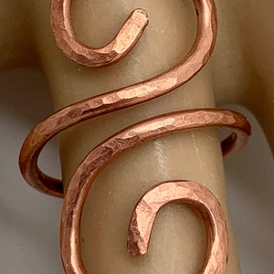 May include: A hammered copper ring with a double swirl design. The ring is made of thick wire and has a rustic, handmade look.