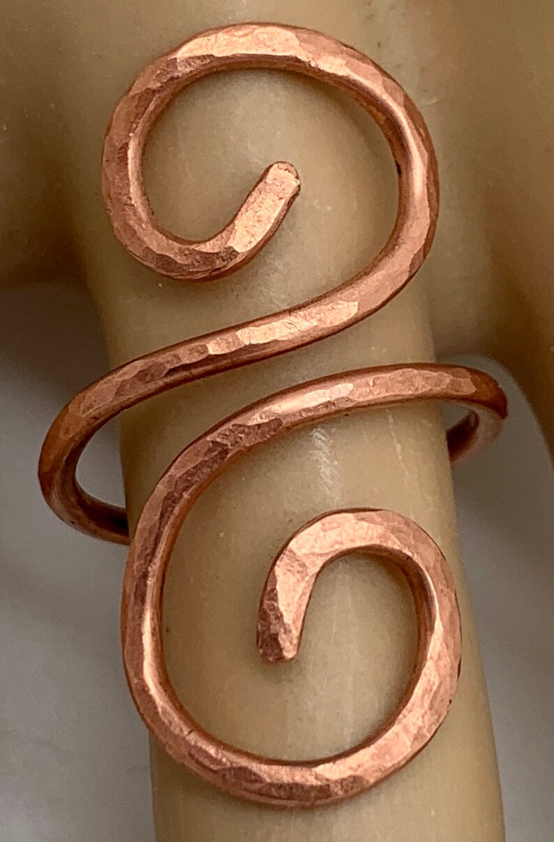 May include: A hammered copper ring with a double swirl design. The ring is made of thick wire and has a rustic, handmade look.