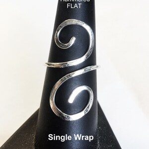 May include: A silver hammered flat ring with a single wrap design. The ring is on a black ring display stand. The text "Hammered FLAT" and "Single Wrap" is visible on the stand.