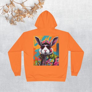 Può includere: Felpa con cappuccio arancione con una grafica colorata di un coniglio con occhiali da sole e un cappello sul retro.