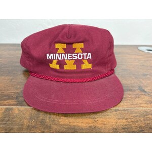 May include: Maroon cap with "MINNESOTA" in white block letters and a gold logo. The cap has a maroon brim and a maroon rope detail. The cap is sitting on a wooden surface.
