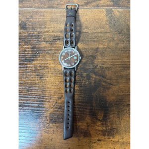 Könnte beinhalten: Eine Armbanduhr mit rundem silberfarbenem Gehäuse und braunem Zifferblatt mit weißen Ziffern und Datumsanzeige. Die Uhr hat ein dunkelbraunes Lederarmband mit einem markanten perforierten Design, das aus zwei Reihen ovaler Aussparungen besteht.