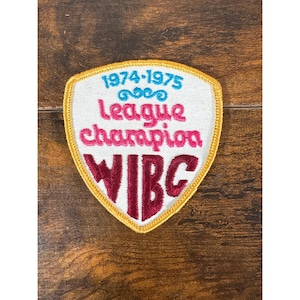 May include: A shield-shaped embroidered patch with a white background and a gold border. It features the light blue text 1974-1975, the pink text League champion, and the dark red text WIBC, all arranged in a stacked design on the fabric surface.