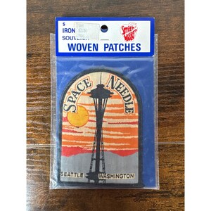 May include: Woven patch featuring the Seattle Space Needle against a sunset backdrop. The patch includes the text "SPACE NEEDLE" and "SEATTLE WASHINGTON". The patch is packaged in a clear plastic bag with a blue header that reads "IRON SOUVENIR WOVEN PATCHES".