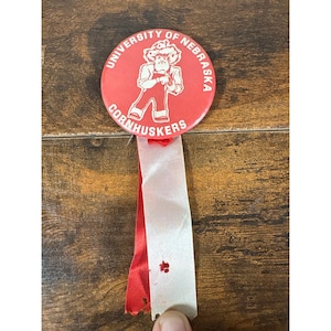 May include: A round red pinback button featuring the white text University of Nebraska Cornhuskers and a white graphic of a mascot. Attached to the bottom are two vertical fabric ribbons, one red and one white, with a small hole in the white ribbon.