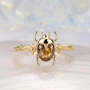 May include: A gold ring featuring a central scarab beetle design with a faceted amber-colored gemstone body. The ring band has a textured, organic appearance resembling a twig or branch, extending from both sides of the beetle centerpiece.