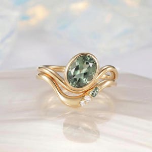May include: A gold ring set featuring an oval-cut green gemstone as the centerpiece. The band has a wave-like design, with smaller green stones and diamonds. The ring is set against a soft, blurred background.