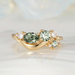 May include: A gold ring featuring a cluster of gemstones. The ring has a flowing design with a central oval green gemstone, surrounded by round green and white gemstones. The ring is set on a white surface.