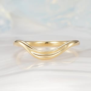 Puede incluir: Un anillo de oro amarillo pulido que presenta una banda lisa y curva en forma de V. La superficie metálica es reflectante y sólida, mostrando un diseño continuo y fluido que se hunde en el centro. El anillo tiene un acabado metálico uniforme.