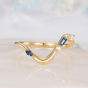 May include: A gold V-shaped ring featuring a marquise-cut blue-purple stone, a rectangular baguette-cut blue stone, and two small round clear stones set along the band. The metal band has a polished finish and a curved, contoured design.