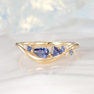 Puede incluir: Un anillo de oro con un diseño ondulado, adornado con zafiros azules y pequeños diamantes blancos. Los zafiros tienen forma de pera y triangular. El anillo está sobre un fondo suave y borroso.