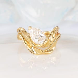 May include: A gold ring set featuring a pear-shaped diamond. The ring has a unique, flowing design with a polished finish. The band curves around the diamond, creating a modern and elegant look. The ring is set on a white surface.