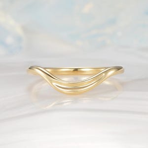 May include: A gold-colored ring with a curved design. The ring has a smooth, polished surface and a wave-like shape. The ring is set against a soft, blurred background.